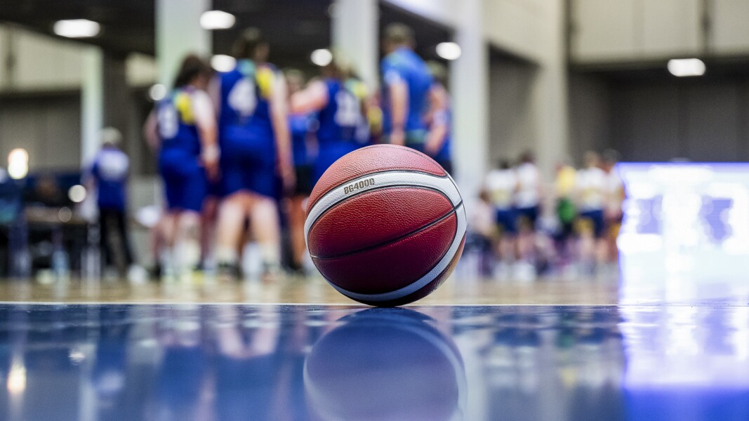 Das Foto zeigt im Fokus einen Basketball, der auf dem Spielfeld liegt. Verschwommen im Hintergrund sieht man die Teams während einer Auszeit.