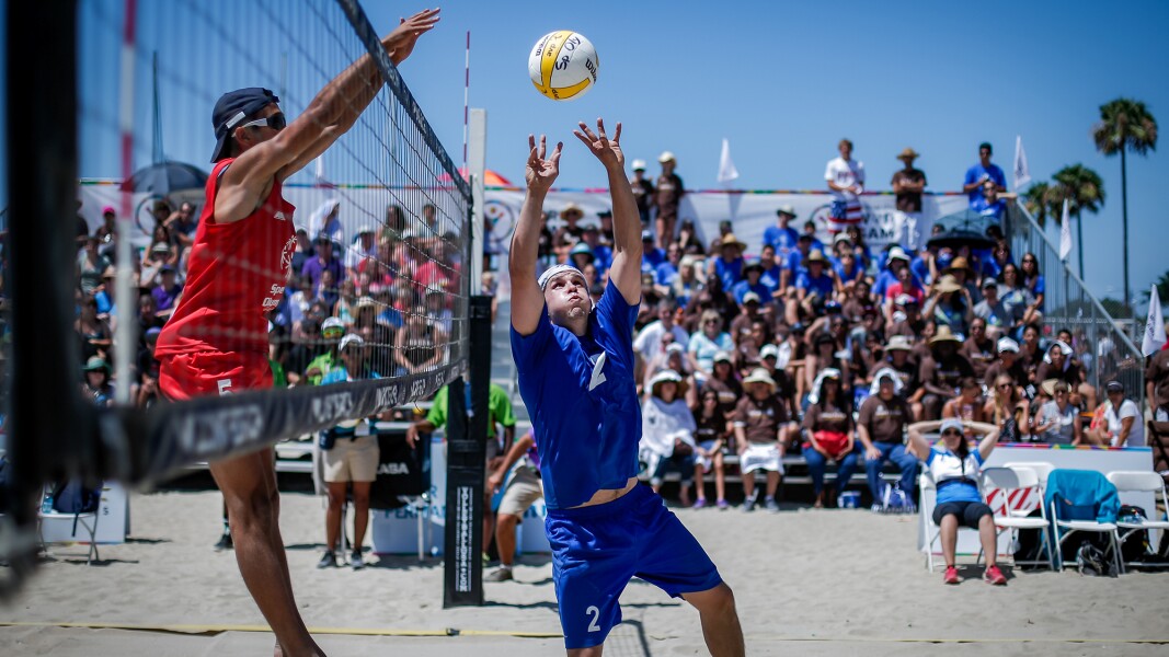 Das Bild zeigt eine Spielszene beim Beachvolleyball. Ein Spiel versucht am Netz zu blocken, während der Gegenspieler pritscht.