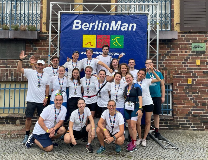 Das Foto ist ein Gruppenbild aller Teilnehmer*innen, die als Teamevent des Weltspieleteams und Special Olympics Deutschland am BerlinMan Triathlon teilgenommen haben. Außerdem dabei war Athlet Marcel Eckardt. Die Gruppe von ca. 18 Personen posiert vor dem BerlinMan Poster.