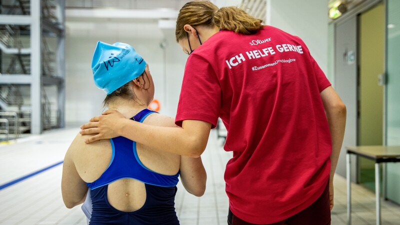 Auf dem Foto sind zwei Menschen von hinten zu sehen. Links ist eine Special Olympics Schwimmerin und rechts ein Volunteer, die ihren Arm um die Athletin gelegt hat. 