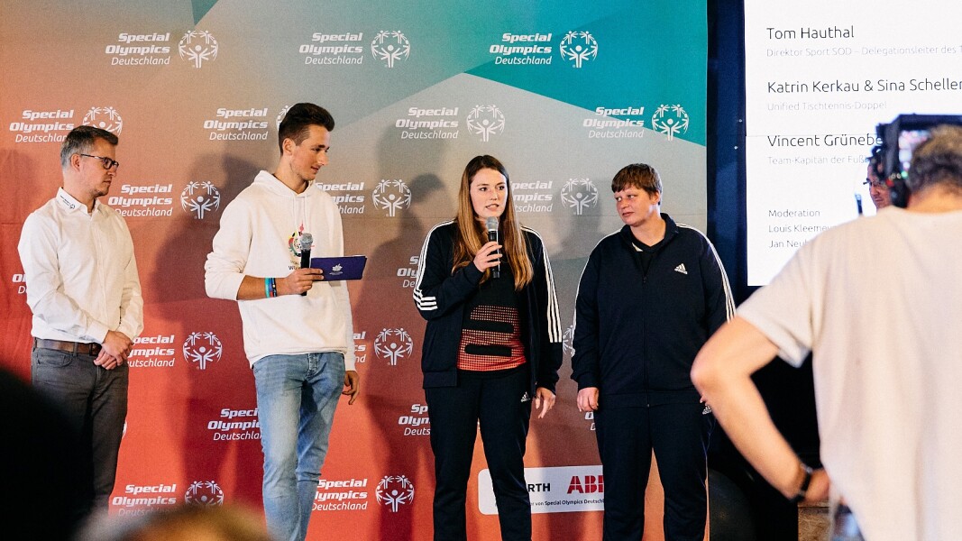 Das Foto zeigt die Bühne mit vier Personen während der Vorstellung des Team SOD für die Weltspiele. Eine Unified Partnerin im Tischtennis spricht gerade ins Mikrofon.