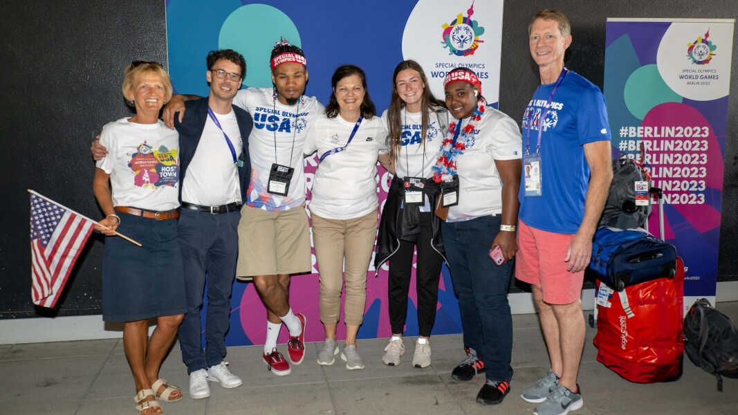 Das Foto zeigt mehrere Mitglieder der US-amerikanischen Delegation nach ihrer Ankunft am Berliner Flughafen. Es sind insgesamt 7 Teilnehmer, die jeweils ihre Arme auf den Schultern ihres Nachbarn haben und lächeln. Ein blonde Frau schwenkt eine kleine amerikanische Flagge.