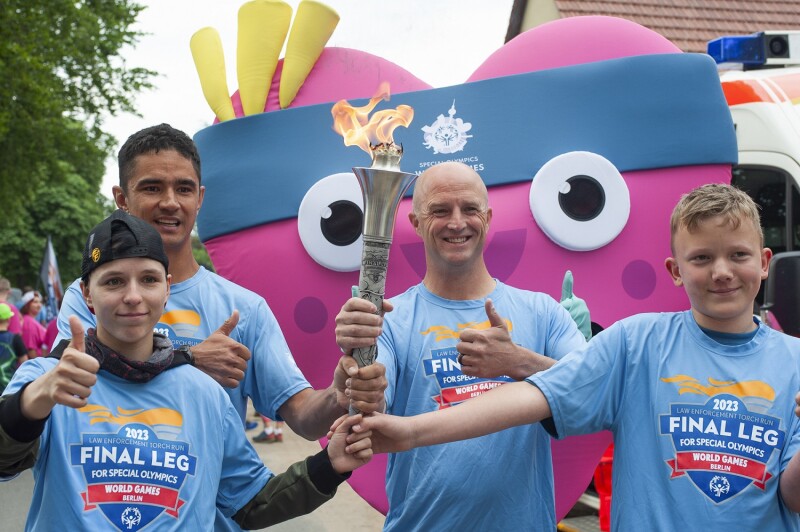 In the photo, the torch is carried by four young kids, who are accompanied by the mascot, a local representative wearing a gray suit, and other people in the background.
