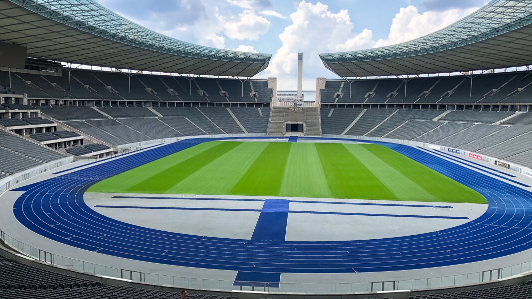 Das Foto zeigt das Berliner Olympiastadion von innen. Es sind Zuschauerränge, Laufbahnen und eine Rasenfläche in der Mitte zu sehen.