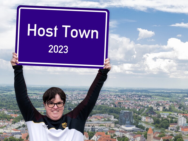 Special Olympics Athletin Juliana Rößler hält ein Schild mit der Aufschrift Host Town in die Luft. Hinter ihr sieht man eine Stadt.