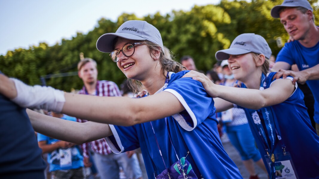 Auf dem Foto sieht man vier Personen. Im Vordergrund sind zwei Mädchen und ein junger Mann zu sehen, die sich an den Schultern halten und eine Polonaise machen. Sie lächeln.