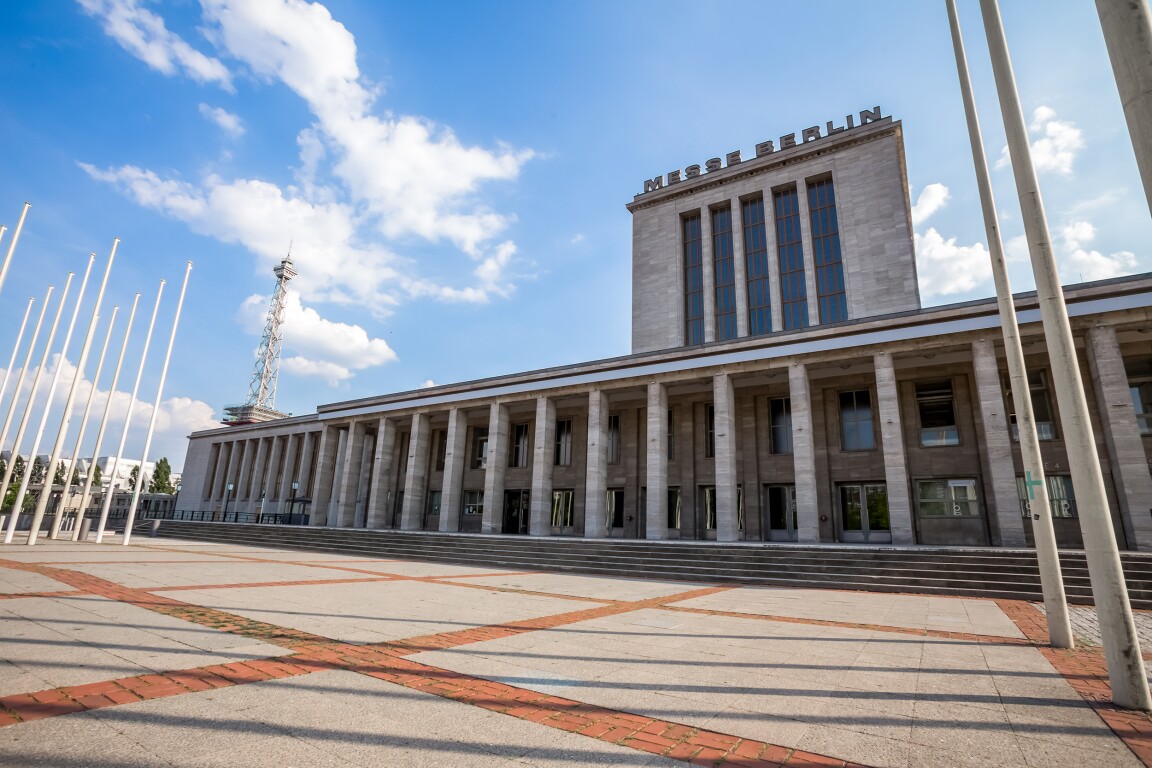 The photo shows a paved forecourt and a building with columns on which Messe Berlin stands.
