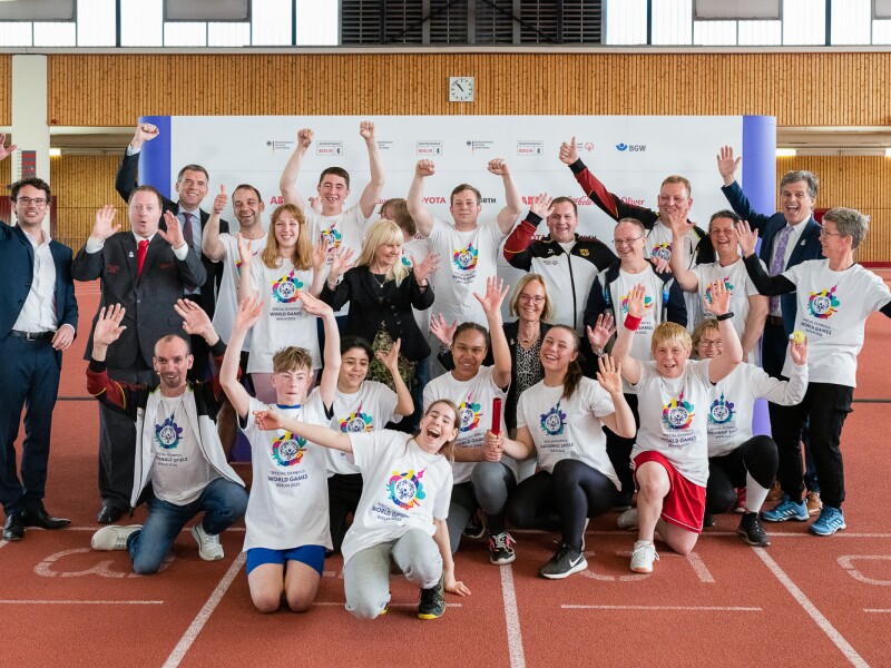 Das Foto zeigt ein Gruppenbild der Pressekonferenz. Alle Athlet*innen, die teilgenommen haben, posieren fröhlich unter anderem mit Sven Albrecht, Timothy Shriver und Ian Harper. Alle haben die Arme nach oben gerissen und jubeln in die Kamera.