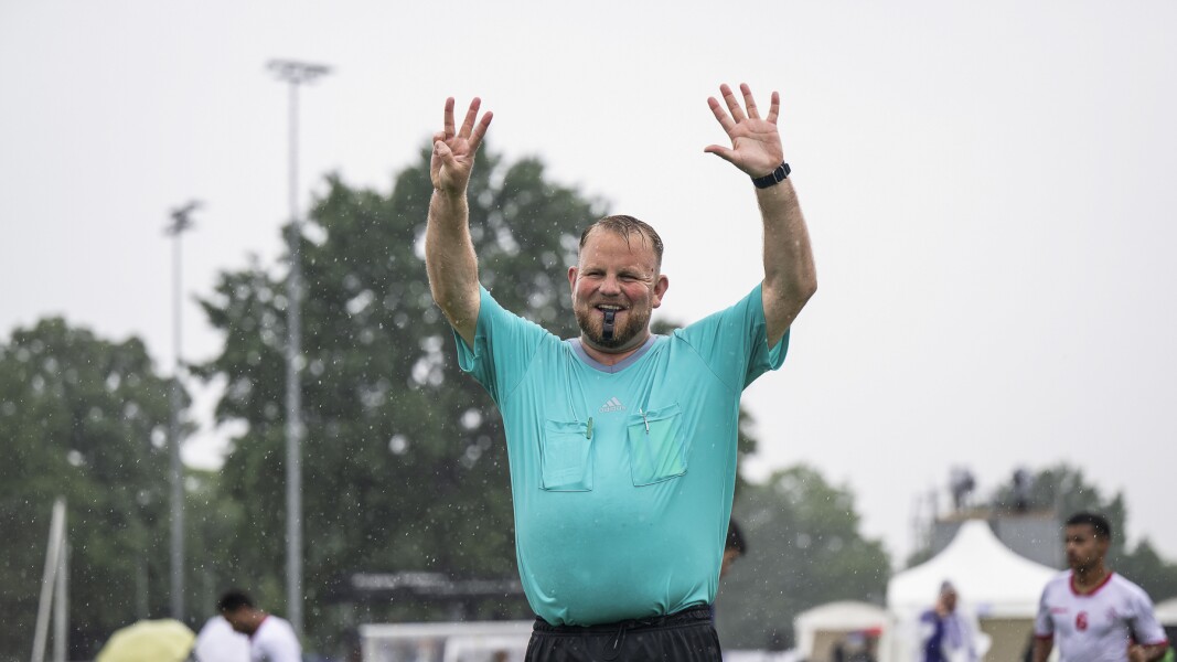 Das Foto zeigt einen Schiedsrichter beim Fußball. Er gibt Handzeichen und lächelt mit seiner Pfeife im Mund. Es regnet, aber er hat trotzdem gute Laune.
