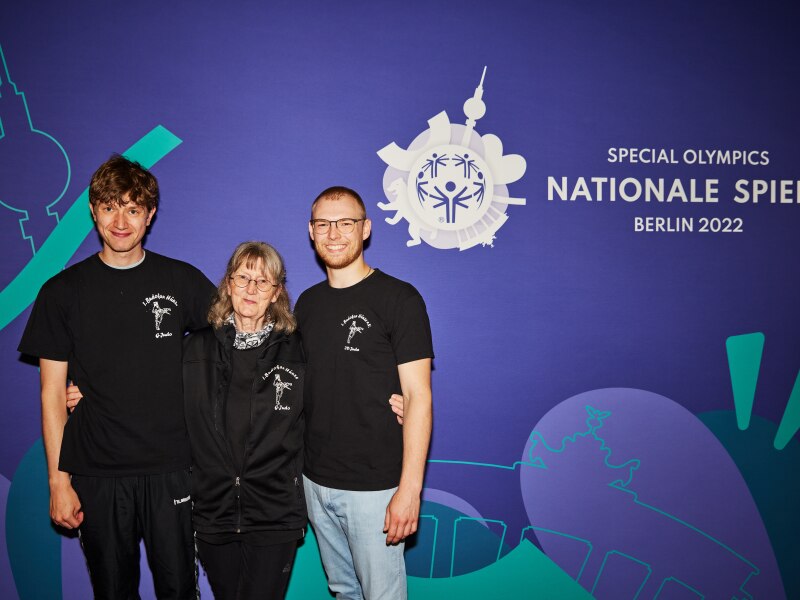 On the photo you see the Unified team together with their coach from left to right: Athlete Thorben von Kneten, coach Gabriele Gramsch and partner Lukas Ehresmann. They stand arm in arm in front of a blue wall on that you see the Special Olympics National Games Berlin 2022 logo.