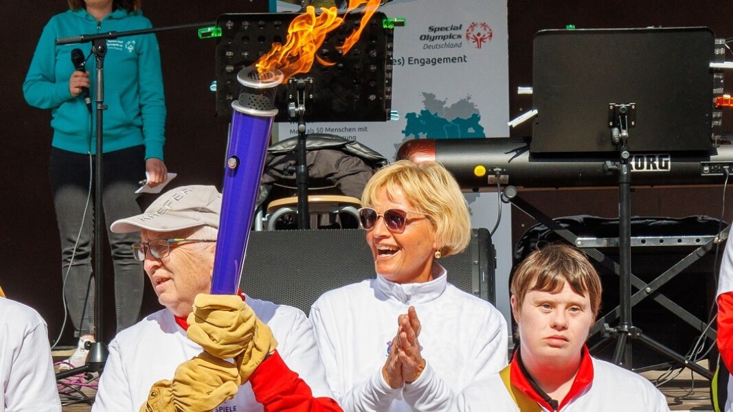 The photo shows a group of people participating in the Bremen Torch Run on the symbolic way to the National Games Berlin 2022. One man is carrying the torch and everyone is clapping their hands. A stage is set up behind the group.