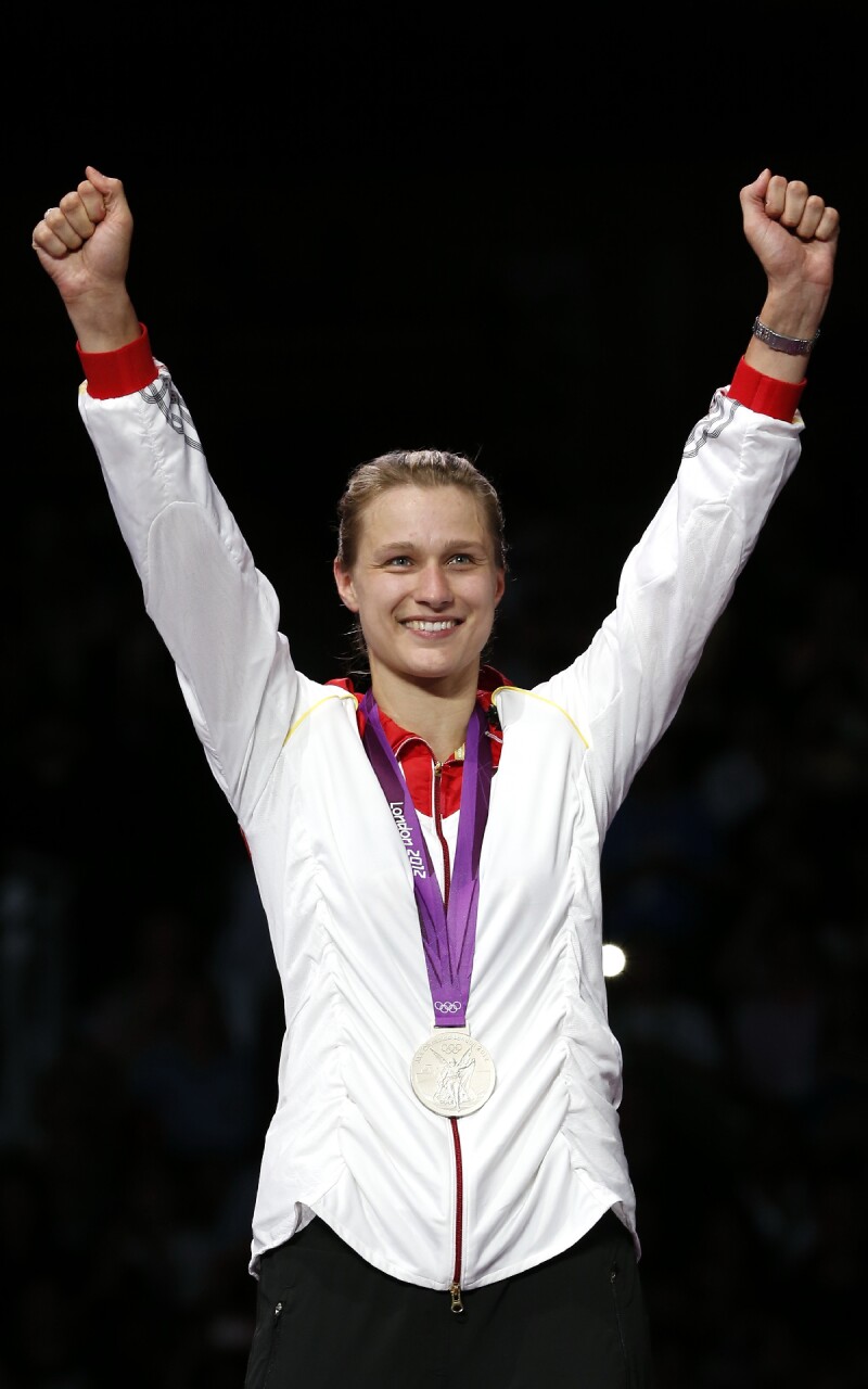 The photo shows the German fencer Britta Heidemann. She has both arms raised in jubilation and is wearing an Olympic silver medal around her neck. 