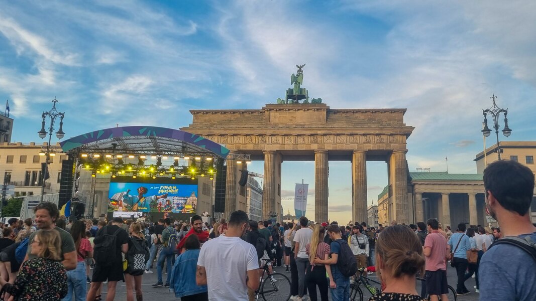 Das Foto zeigt eine belebte Szene vor dem Brandenburger Tor. Die Bühne in Kooperation mit der Fete de la Musique ist direkt davor aufgebaut und viele Menschen tummeln sich davor.