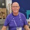 The picture shows Volunteer Bernd sitting and smiling in his purple uniform at the volunteer materials distribution booth.