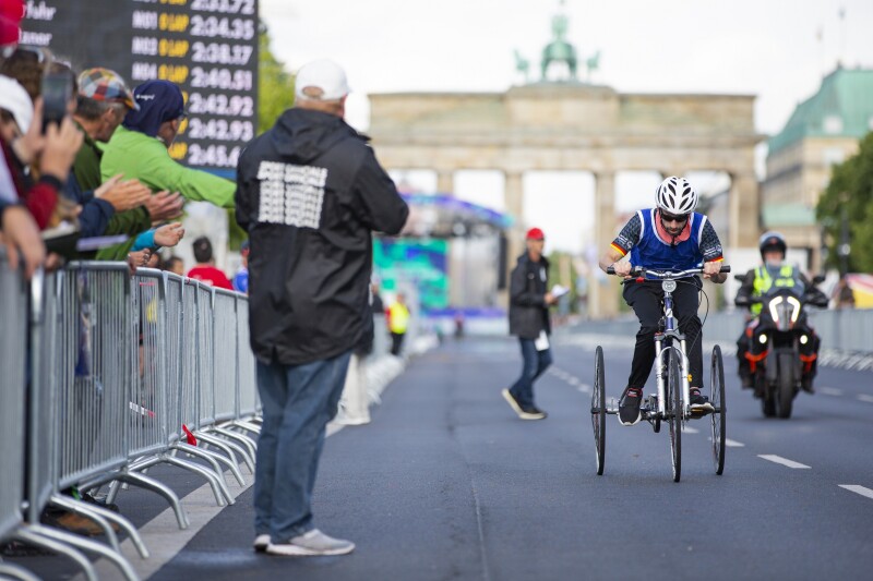 The photo shows cyclist and one of the faces of the Games Robert Herberg in action during a bike race. The Brandenburg Gate can be seen in the background and many spectators are cheering on the left side.