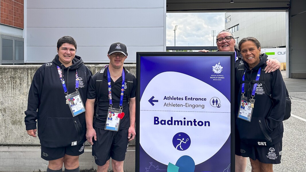 Auf dem Foto sind Athlet*innen vor dem Badminton Schild zu sehen. Sie schauen glücklich