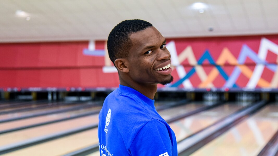 Auf dem Bild lacht ein Bowling-Spieler in die Kamera. Im Hintergrund sind die Bowling-Bahnen.