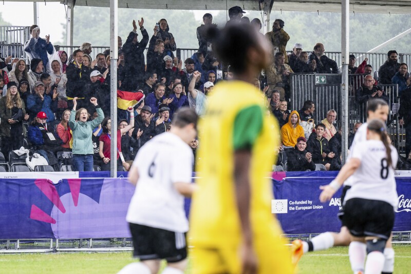 Das Foto zeigt eine Szene beim Fußball. Die Zuschauer*innen jubeln über ein Tor des deutschen Teams, das trotz starkem Regen eine super Leistung zeigt. 