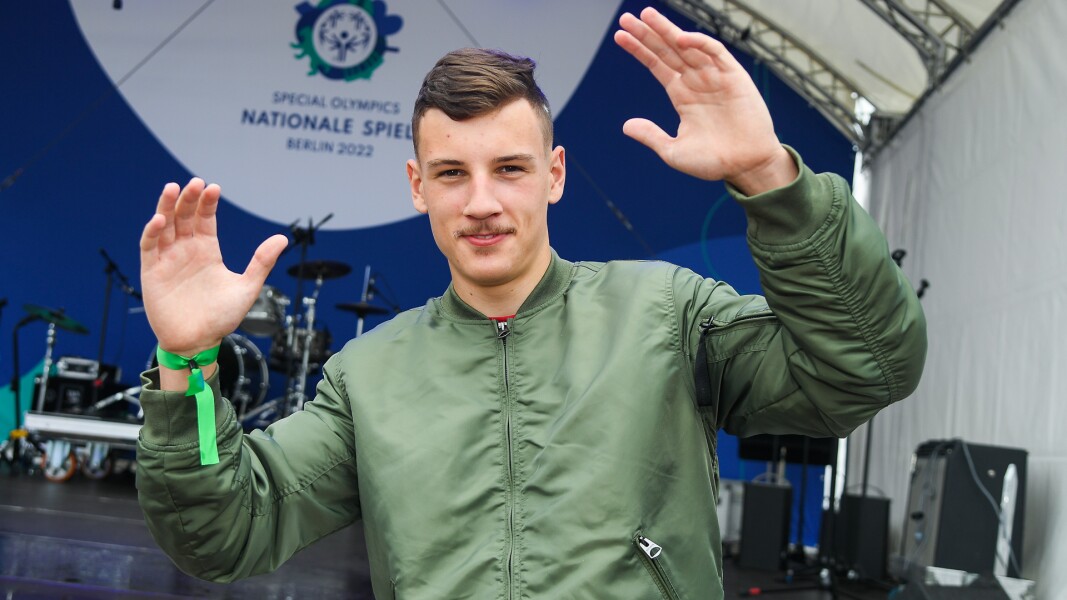 Auf dem Foto sieht man Rapper Pusha CL vor der Bühne auf dem Special Olympics Festival. Er trägt eine grüne Jacke, hält beide Arme nach oben und lächelt.