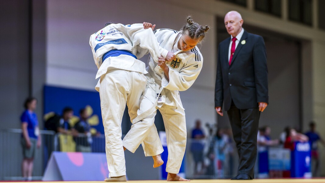 Zwei  Judo-Kämpferinnen ringen miteinander unter Beobachtung des Schiedsrichters.