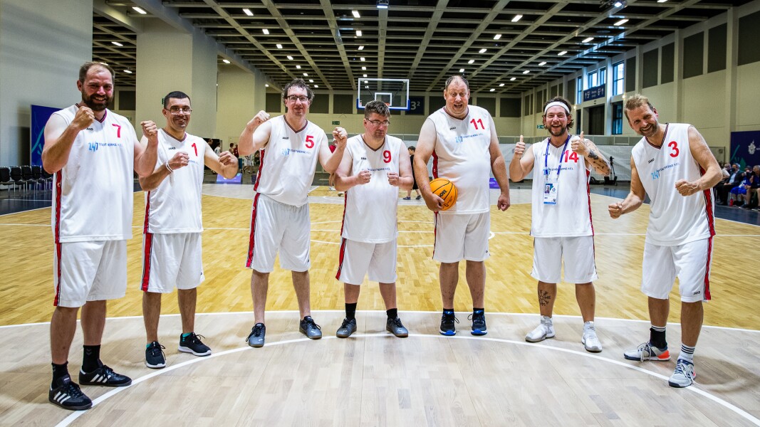Auf dem Foto steht ein Basketball-Team im Halbkreis auf dem Spielfeld. Alle jubeln freudig in die Kamera und ein Spieler hält einen Basketball. Es ist keine Spielsituation.