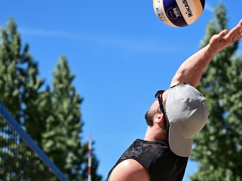 The photo shows a player in action at a beach volleyball competition. He is about to execute an attacking shot. He is wearing a cap and sunglasses.