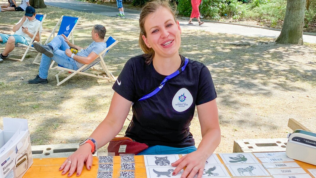 Das Bild zeigt Christin Hertwig, die einen Stand beim Special Olympics Festival betreut. Sie sitzt an ihrem Stand und lächelt in die Kamera.
