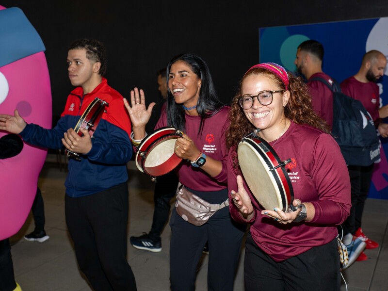 Das Bild zeigt die Ankunft der Delegation aus Puerto Rico. 3 Sportler*innen lächeln, singen und tanzen mit einem Tamburin in der Hand.