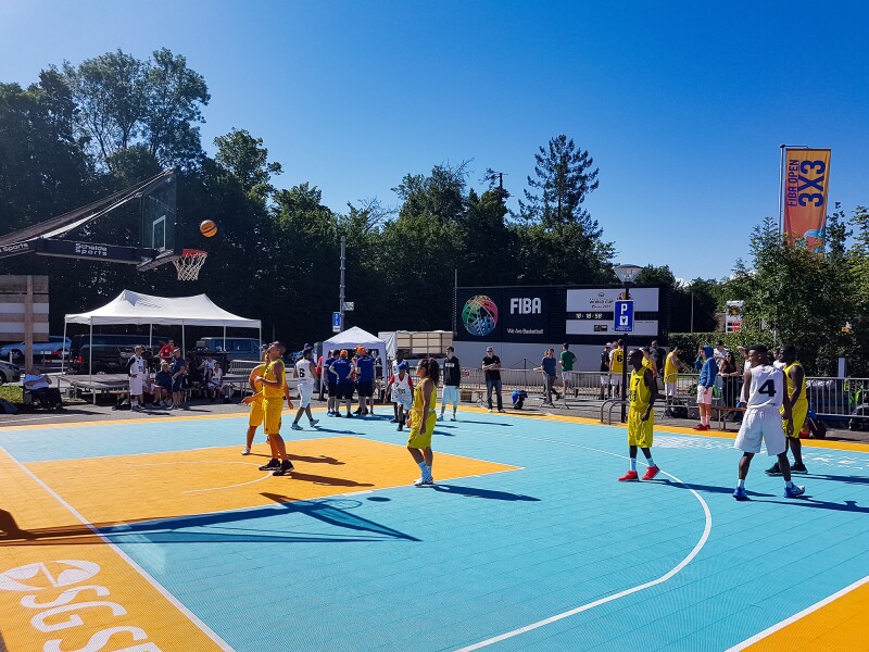 The photo shows a 3x3 Basketball pitch outside, with two teams preparing for competition. 