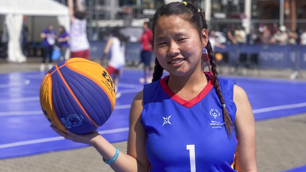 In the picture, a female basketball player is smiling at the camera. She holds a basketball in one hand and stands in front of the 3x3 court.
