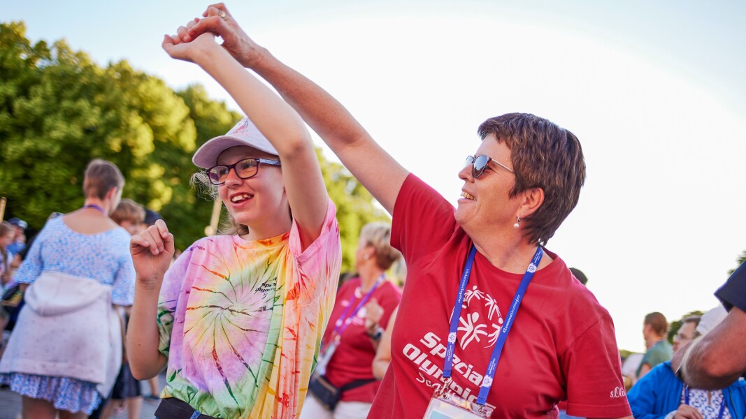 Auf dem Foto tanzen zwei Frauen, eine jüngere Athletin gemeinsam mit einer Frau, die ein rotes T-shirt mit Special Olympics drauf trägt. Beide lächeln und freuen sich.