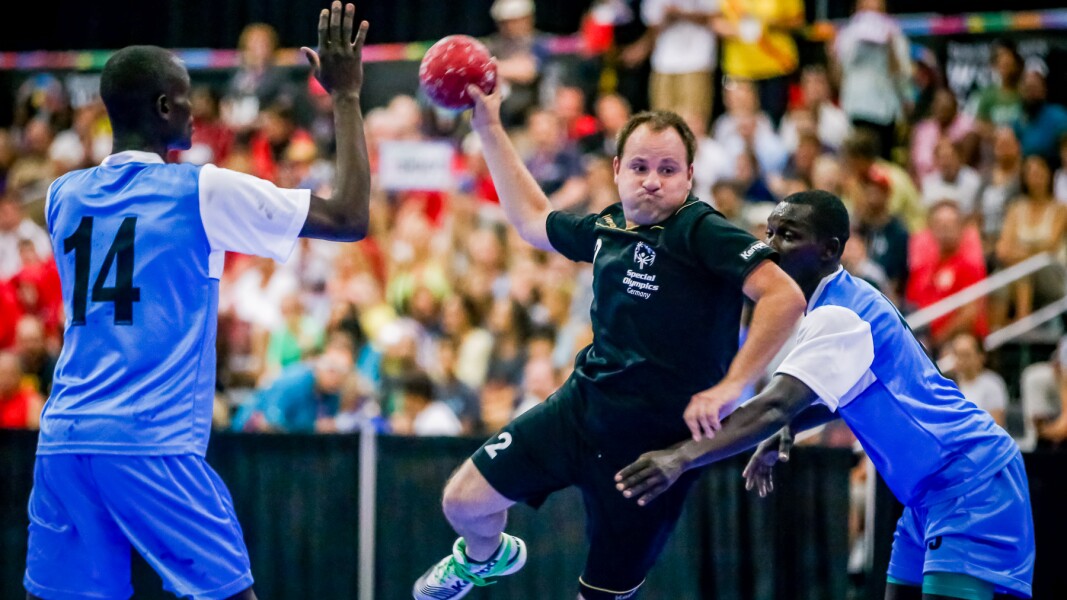Das Foto ist während eines Special Olympics Handballspiels aufgenommen. Im Vordergrund ist ein Athlet im Sprungwurf und zwei gegnerische Spieler versuchen ihn zu verteidigen. Im Hintergrund sind volle Zuschauerränge.