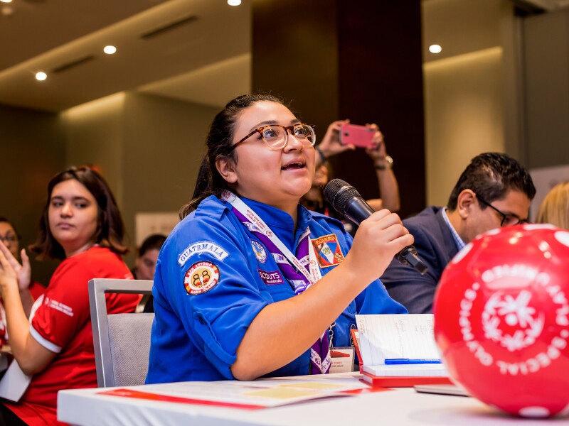 Das Foto zeigt eine Special Olympics Botschafterin mit einem Mikrofon. Sie spricht und alle hören ihr zu. Vor ihr liegt ein Fußball.