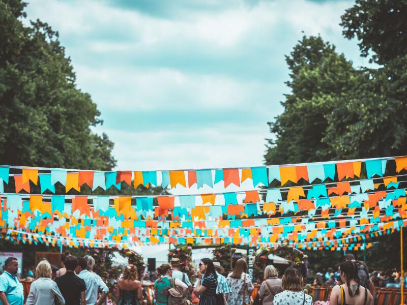 Das Foto zeigt eine Festival-Szene. Viele bunte Wimpelketten sind über den Menschen zwischen zwei Baumreihen gespannt.