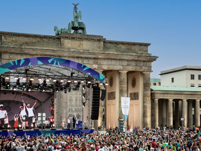 In the photo you can see the panorama of the athletes' disco at the Brandenburg Gate. In front of it is a big crowd and everyone is having fun and raising their arms.