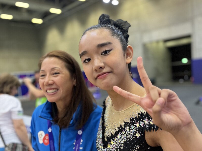 There are two people in the photo: A rhythmic gymnastics athlete is showing "peace" and the woman next to her is laughing happily.
