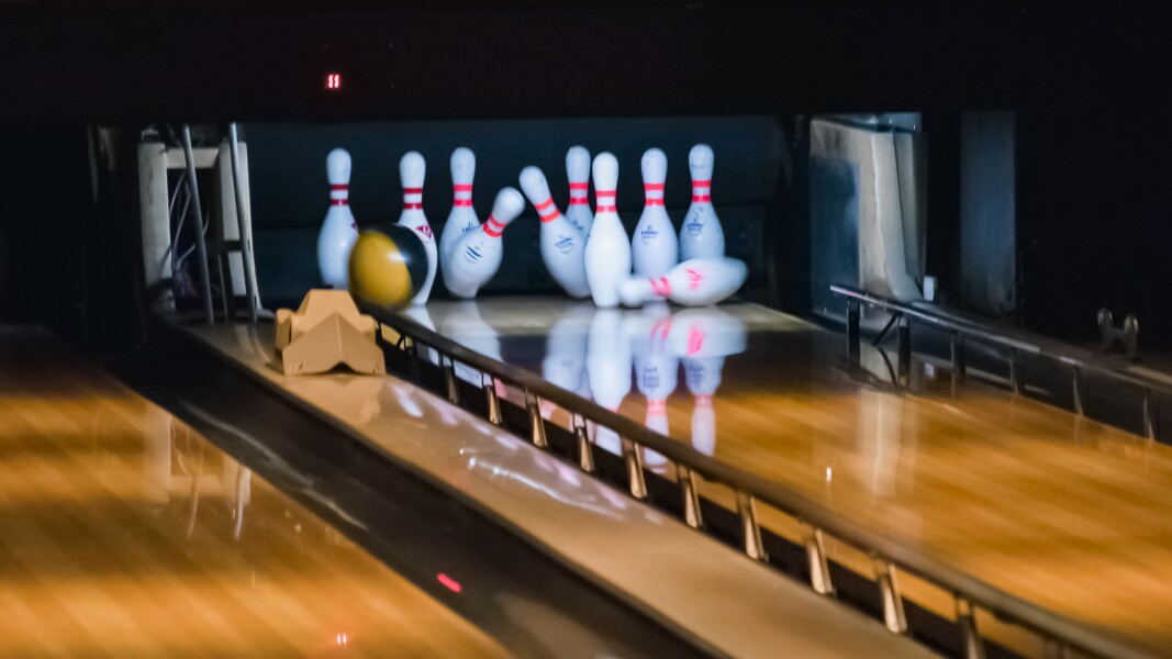 Das Foto zeigt das Ende einer Bowlingbahn und eine Bowlingkugel die gerade Kegel umwirft.