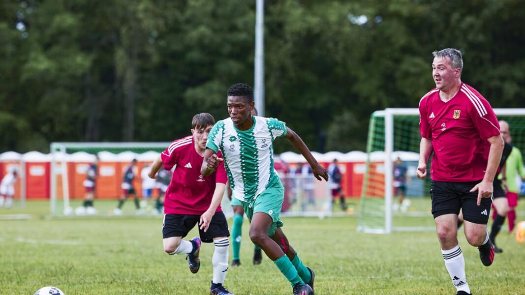 South African football player in green and while jersey getting close to the ball while being chased by two German football players in red jerseys and black shorts.