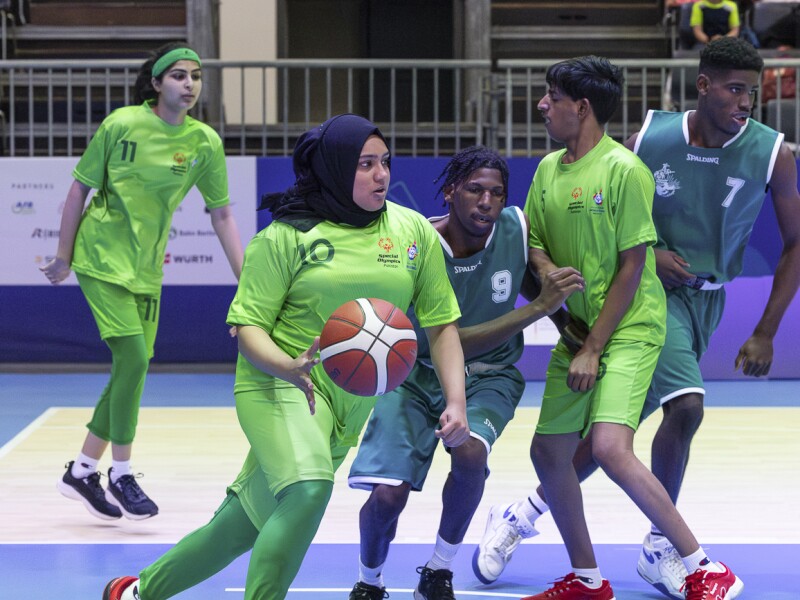 Im Bild fünf Basketballspielerinnen, von denen eine Spielerin den Ball führt und Unterstützung durch Blocken herhält.