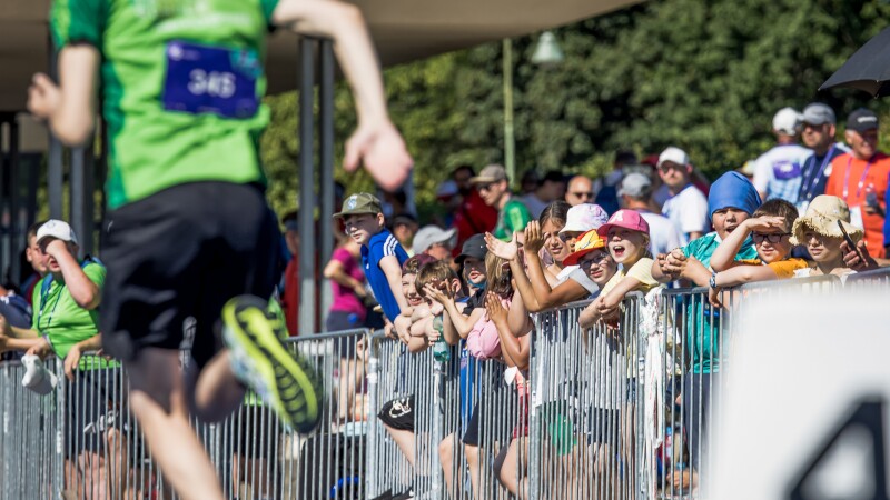 In the foreground of the photo is a track and field athlete sprinting, in the background is a group of young people cheering. They participated in the Fans in the Stands program. 