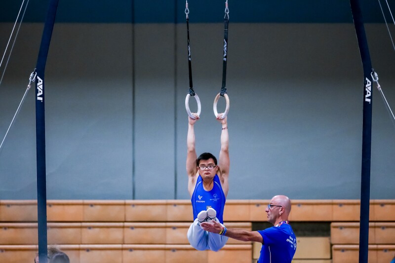 The photo shows a male gymnast at the rings.