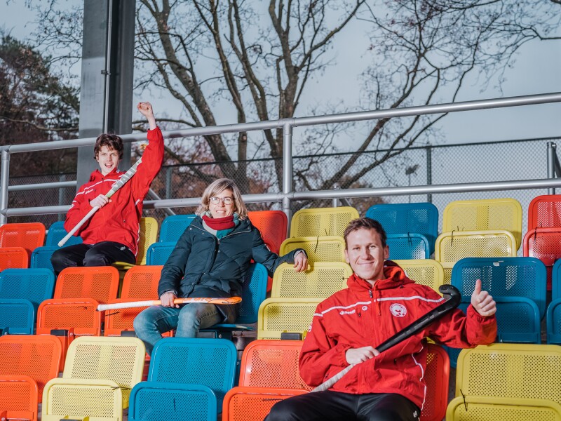 The photo shows three people holding field hockey sticks and sitting on a stand. All are smiling.