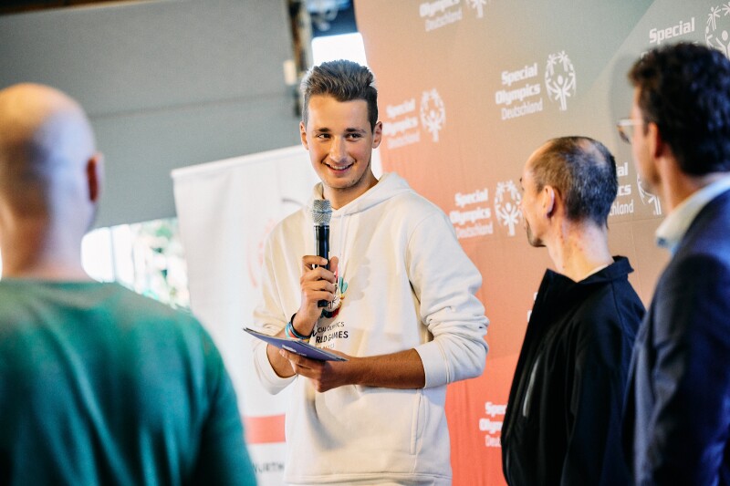 Auf dem Foto ist Louis mit einem Mikrophone zu sehen, er spricht zu anderen Menschen und lacht dabei.