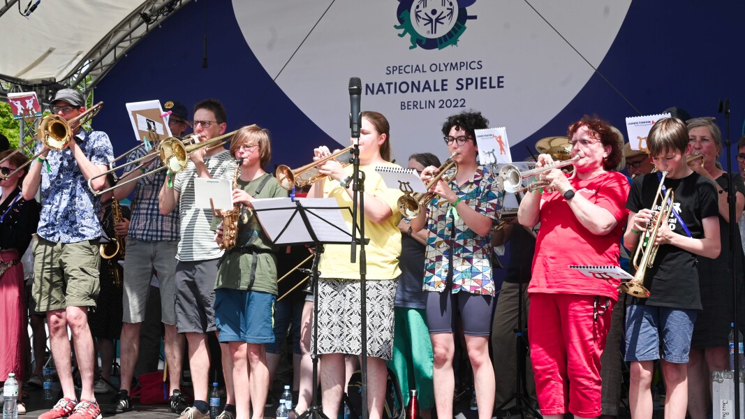 On the photo you can see many musicians of the band Grand Fafare. They are standing on a stage and playing a piece with their wind instruments.