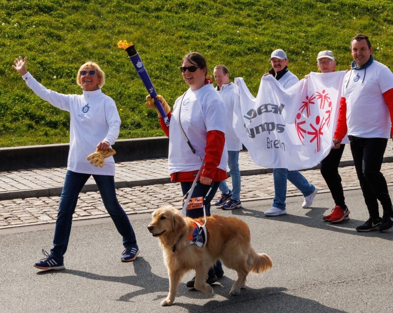 Das Foto zeigt eine Szene des Bremer Fackellaufes auf dem symbolischen Weg zu den Special Olympics Nationalen Spielen Berlin 2022. Es sind insgesamt 6 Menschen und ein Begleithund zu sehen. Die glücklich lächelnden Personen tragen eine Special Olympics Fahne und eine Athletin trägt die Fackel.