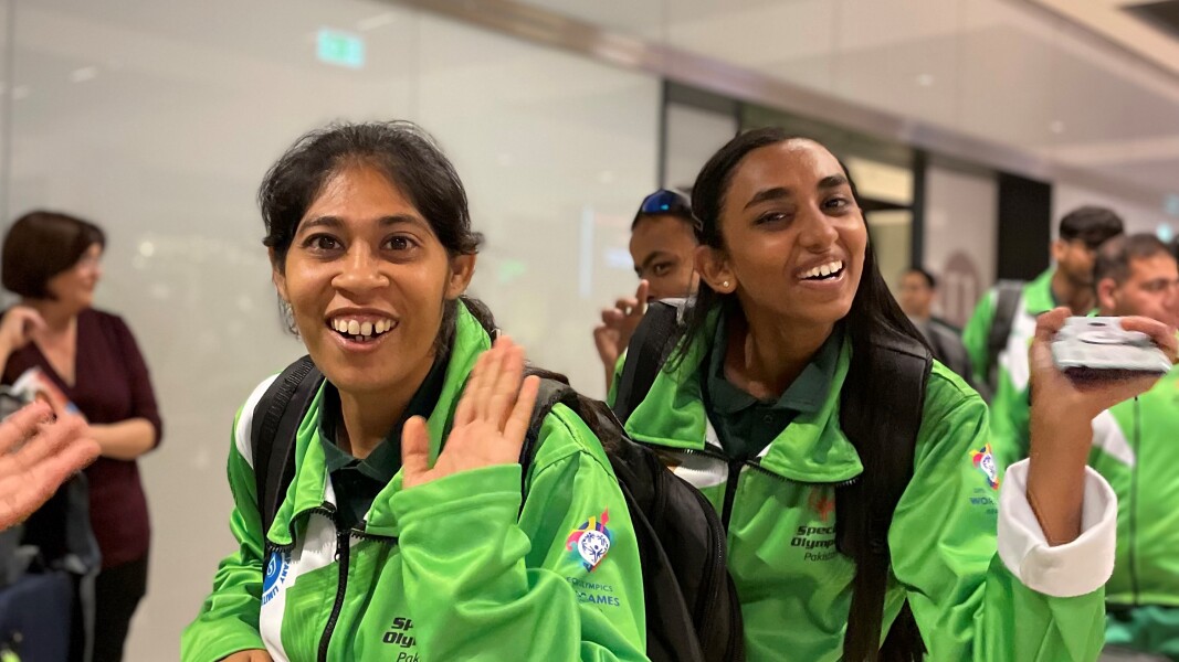 Das Foto zeigt die Ankunft der Delegation aus Pakistan zu den World Games Berlin 2023 am Flughafen Berlin. Vorn im Bild sind 2 junge Frauen mit grünen Trainingsanzügen zu sehen, die fröhlich in die Kamera lächeln.