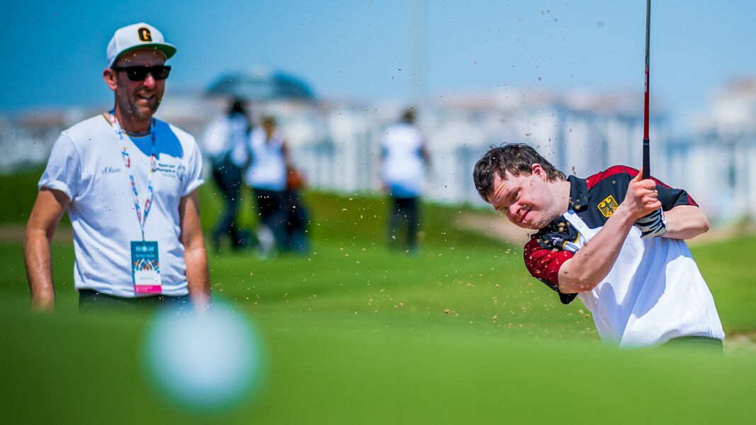 Auf dem Bild ist ein Golfspieler in Aktion zu sehen. Sand fliegt von seinem Schlag durch die Luft.
