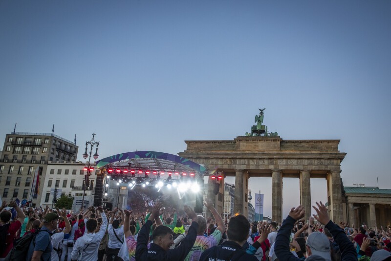 Auf dem Foto ist das Panorama der Athletendisco am Brandenburger Tor zu sehen. Davor ist eine große Menschenmenge und alle haben Spaß und die Arme nach oben gerissen.