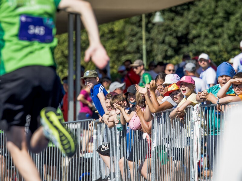 In the foreground of the photo is a track and field athlete sprinting, in the background is a group of young people cheering. They participated in the Fans in the Stands program.