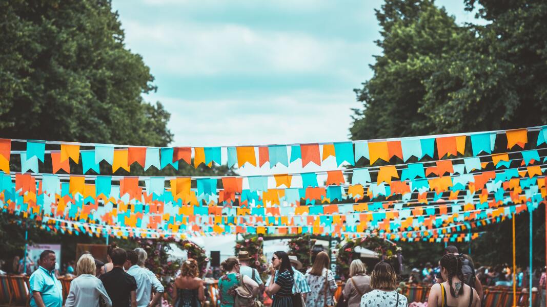 Das Foto zeigt eine Festival-Szene. Viele bunte Wimpelketten sind über den Menschen zwischen zwei Baumreihen gespannt.